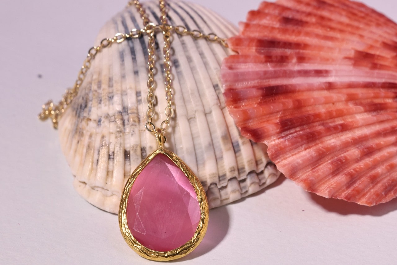 a yellow gold pendant necklace with a pink stone lying on shells