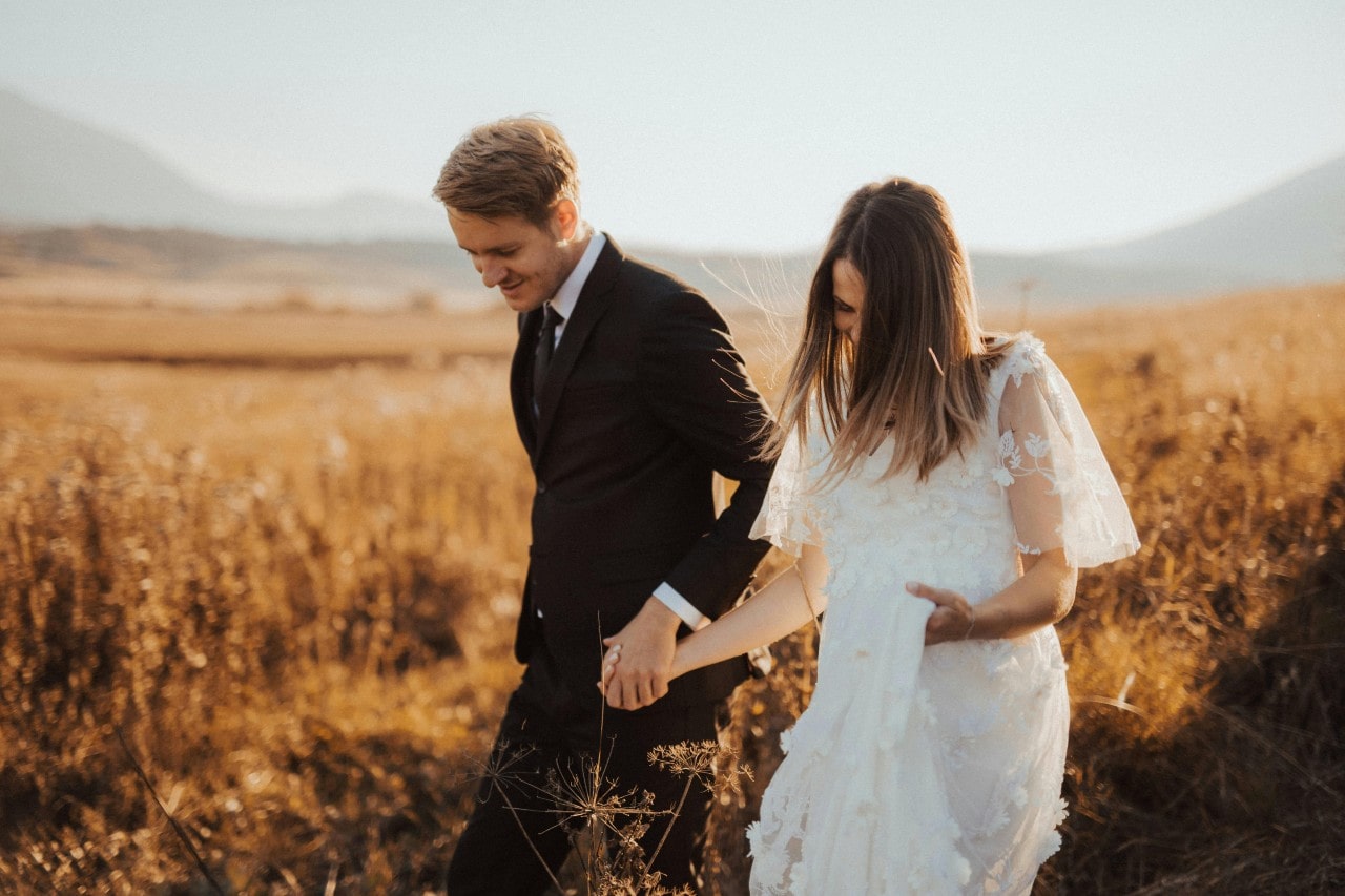 a bride and groom walking hand-in-hand through a field in the fall