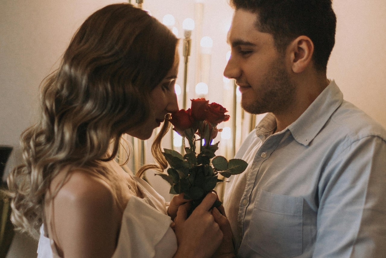 A couple spending Valentine’s Day together; the man is holding red roses the woman is then smelling