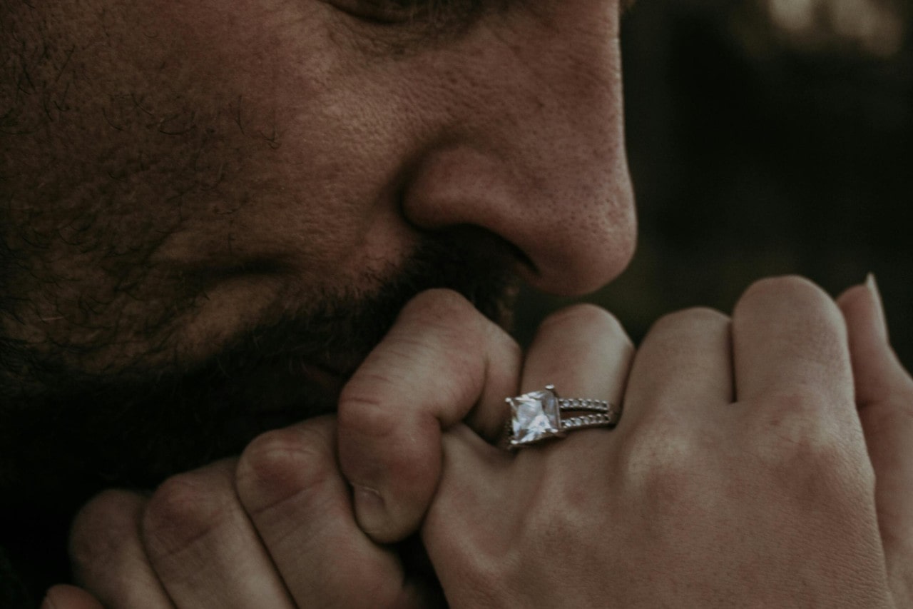 A man kissing a woman’s hand that is adorned with a princess cut engagement ring.