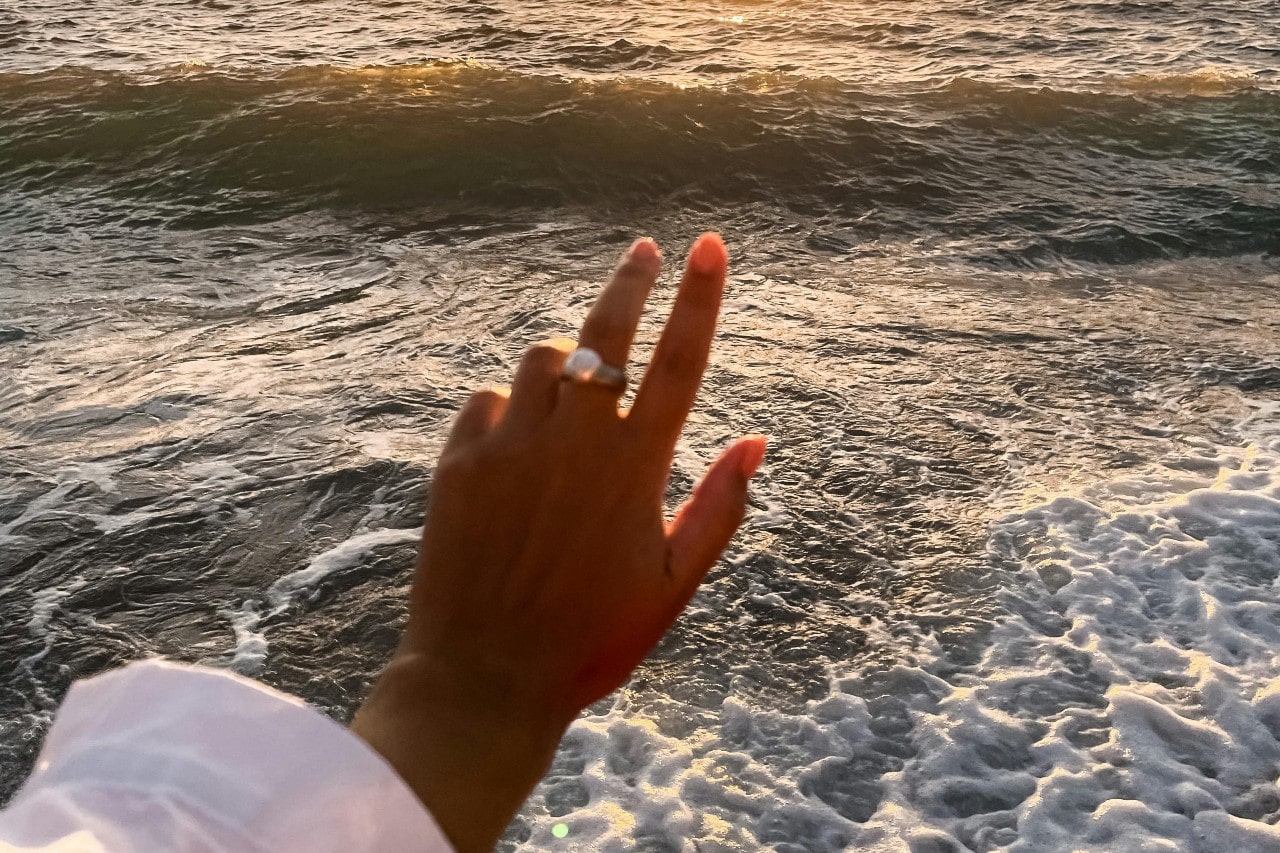 a woman’s hand extended toward the ocean, wearing a large pearl ring.