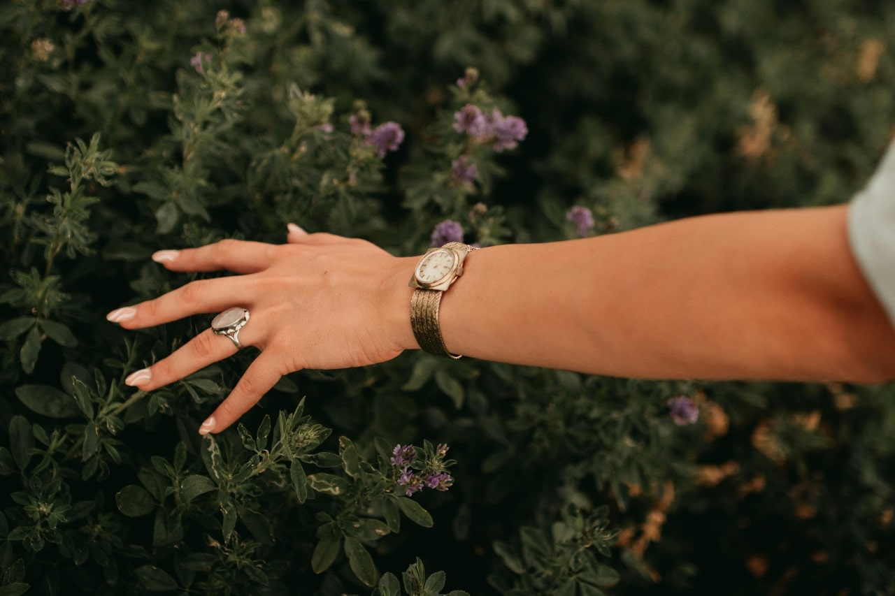 A woman’s hand touching a flower bush, wearing a statement ring and gold watch.