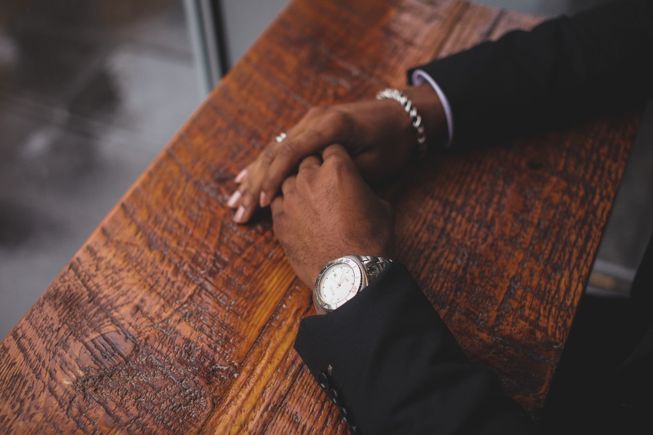 A close-up of a well-dressed man’s hands atop a wooden counter, adorned with a luxury watch, a chain bracelet, and a matching ring.