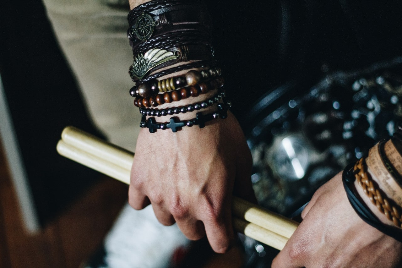 A close-up of a man’s hands holding a pair of drumsticks, adorned in multiple bracelets.